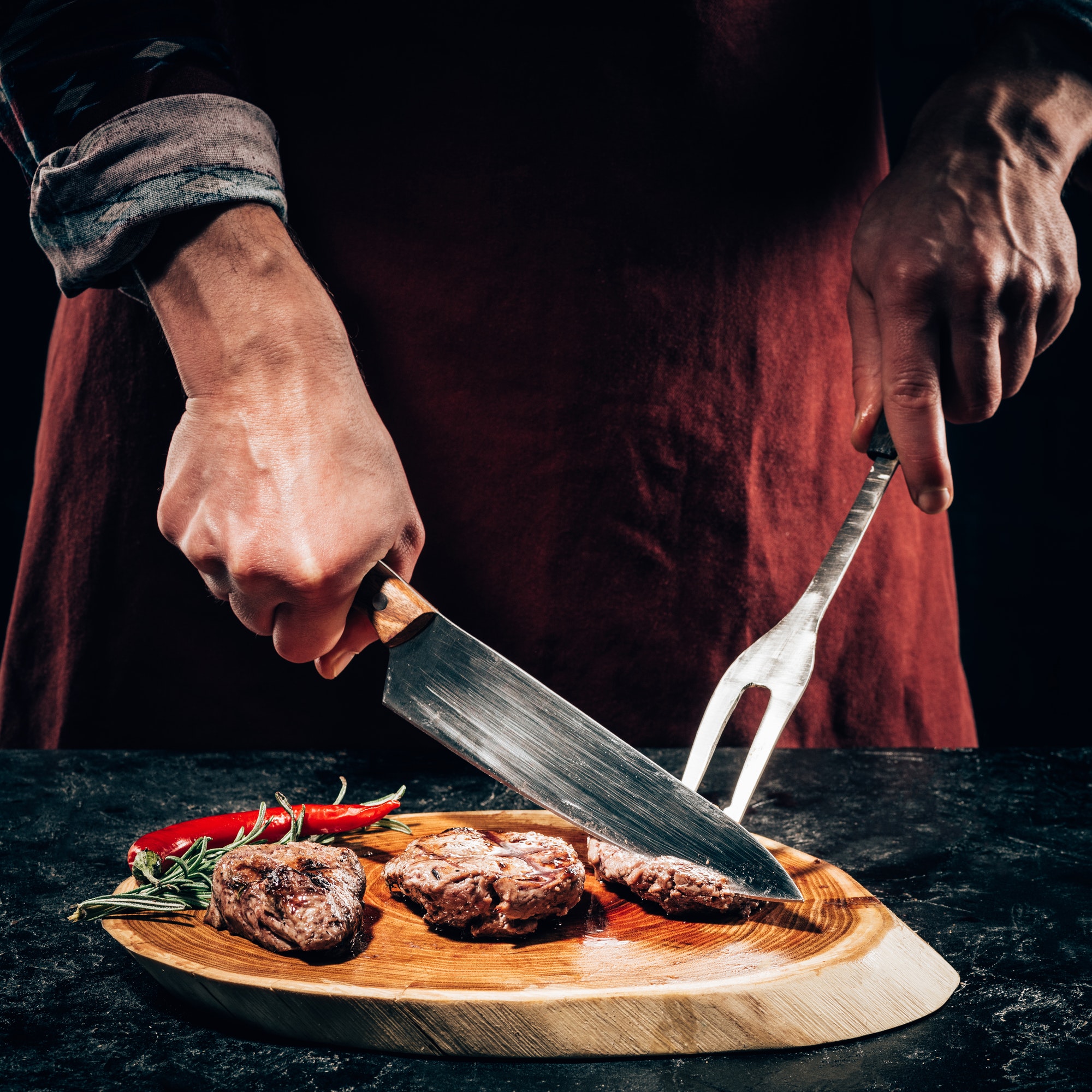 close-up partial view chef in apron with meat fork and knife slicing gourmet grilled steaks with